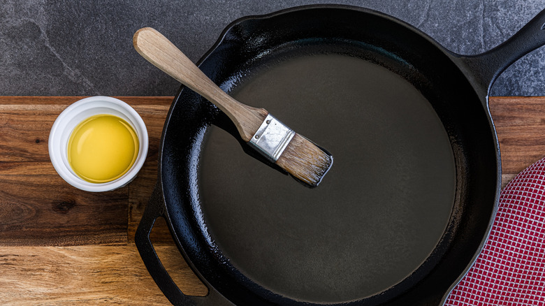 A pastry brush sits in a well-oiled cast iron pan. There is a small bowl of oil next to the pan.