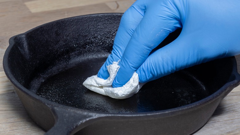 A hand wearing a blur latex glove is using a paper towel to oil a cast iron skillet.