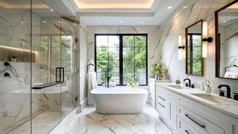 large primary bathroom with marble-look veined tile walls and floor, a freestanding tub under a large back window, a walk-in shower behind glass, and a double white vanity