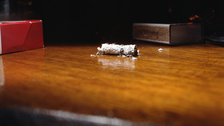 cigarette ash resting on wooden  table with a box of matches and deck of cards in the background