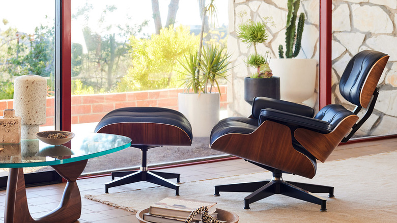 replica black leather and wood Eames lounge chair and ottoman in front of a large glass window with potted succulents outside