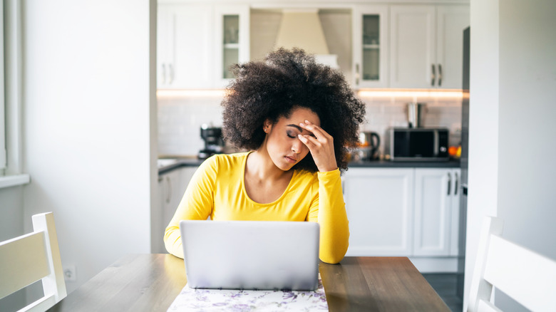 A stressed out woman sitting at a dining table with her laptop with a white kitchen with white walls and subway tile in the background