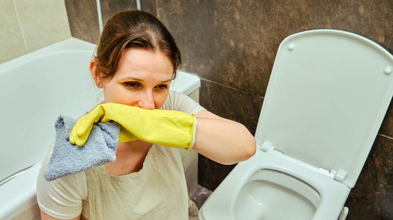 White woman wearing yellow gloves holding blue rag holding back gross smell while cleaning toilet in disgust