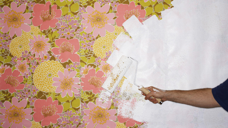 someone using a roller to apply white paint over pink and yellow floral wallpaper