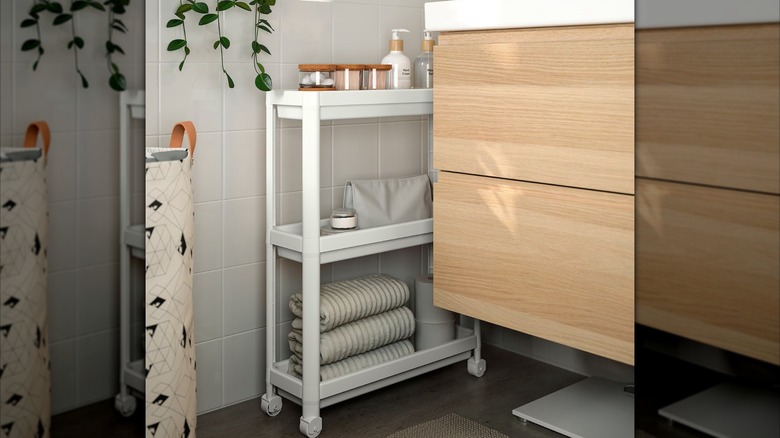 white IKEA VASKEN cart with extra bathroom towels, products, and toilet paper rolled part way out in a narrow space between the wood bathroom vanity and white tiled wall