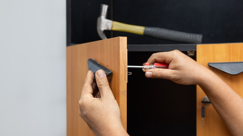 A man's hands are screwing on a cabinet door pull with a hammer in the background.