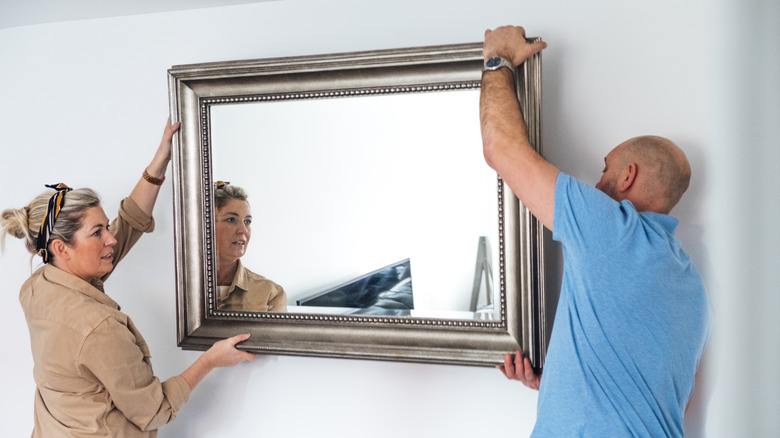 A middle-aged man and woman are hanging a large mirror with a silver frame on a wall.