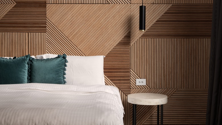 A bedroom with a wood-textured wall. Each wood panel is carved with lines in a variety of patterns. There is a bed dressed in white with 2 green pillows and a minimalist nightstand.