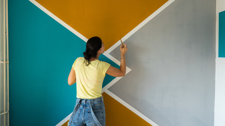 A woman wearing a yellow t-shirt and overalls is painting a wall. She is painting a triangle pattern in shades of teal, gold, and gray.