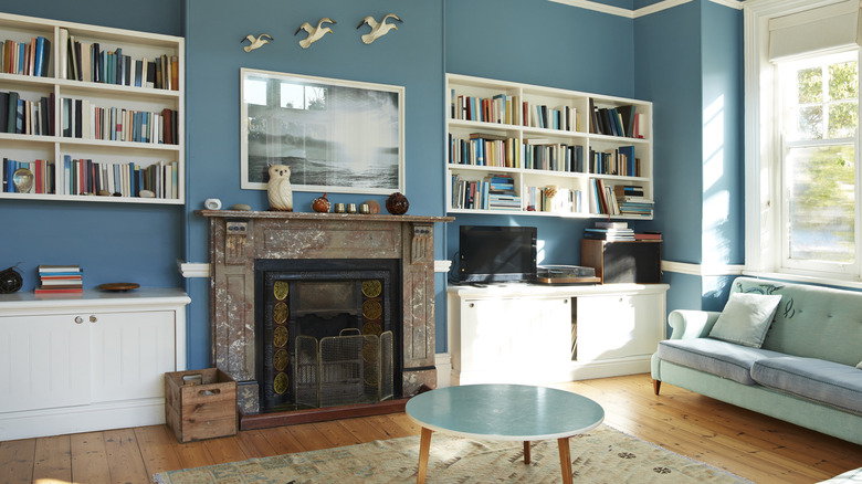 A living room is painted a dark turquoise. There are built-ins on either side of the fireplace and a sofa under the large window.