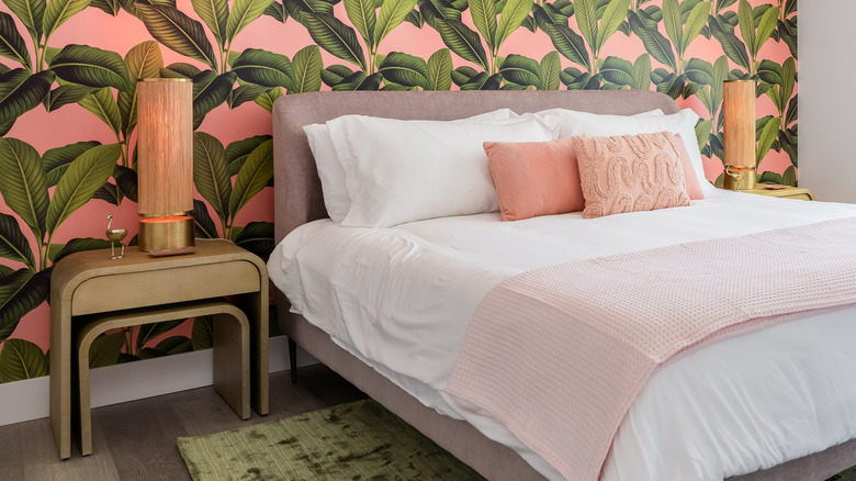 Pink wallpaper covered in a tropical banana leaf print covers the wall behind a bed. The bed is dressed in pink and white. Matching gold nightstands with pink and gold lamps are on either side.