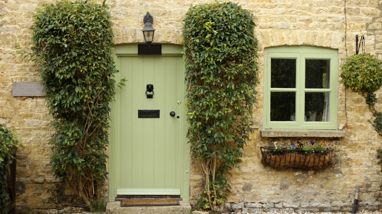 The entryway of an old stone home, which looks straight out of the French countryside. There are two large topiaries surrounding the sage green front door, and a window box of flowers under the sage green window.