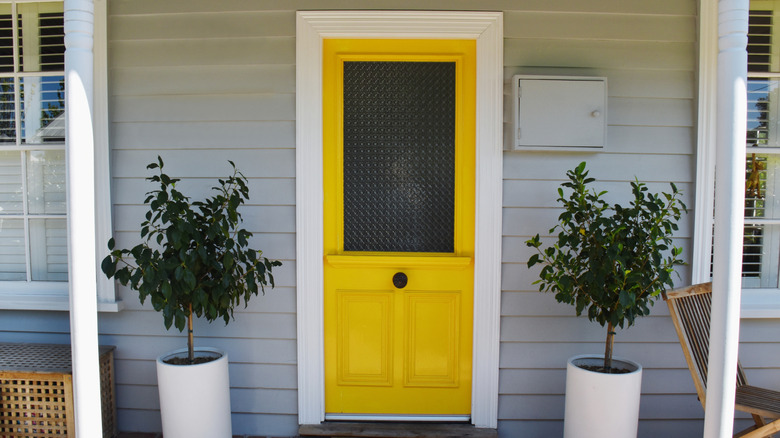 The front porch of a home with light gray siding, white trim, and a bright yellow door. There are white pots of trees on either side of the door.