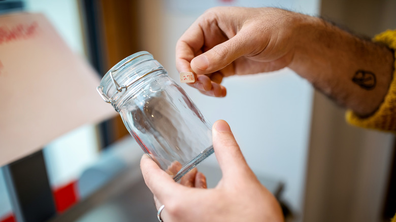 A man's hand is placing a price tag sticker on a glass jar.