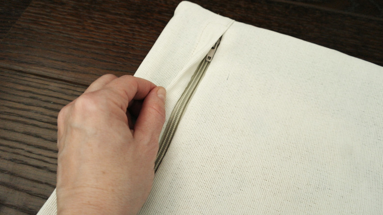 An off-white pillow cover sits on a dark wood table. A man's hand is pulling back the upper flap to reveal the zipper.