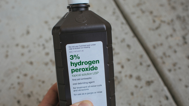 hand holding a brown bottle of 3% hydrogen peroxide. The background is a slightly blurred, flecked floor.