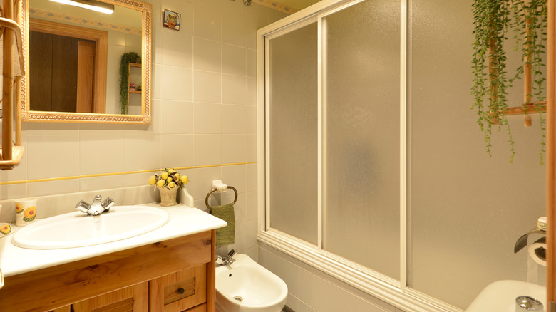 An older bathroom with wood features includes a framed glass shower door with frosted glass.