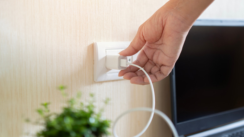 A woman's hand is plugging in a phone charger into the wall outlet. There is a television in the background.