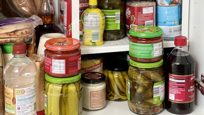 blonde woman staring at camera while she puts away grocery items into a crowded pantry cabinet