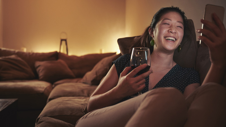 A smiling young Asian woman is sitting on a sectional with a glass of red wine while scrolling through her phone. The room is dimly lit with soft lighting behind the sectional.