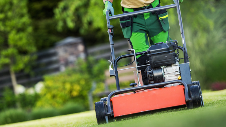 A landscaper is shown from the waist-down mowing a short, green lawn.