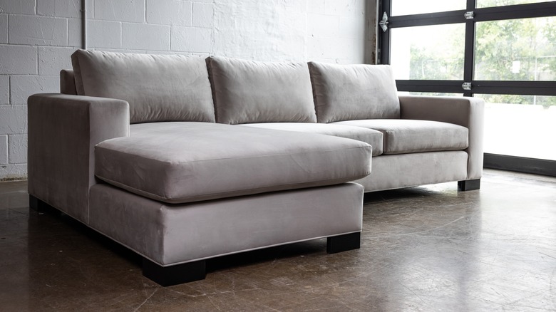 gray velvet sofa with large chaise seat in an industrial living space with white cinderblock walls, concrete floors, and a closed black overhead door of glass.