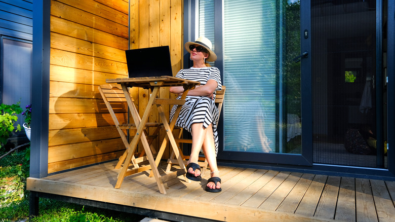 woman wearing hat and sunglasses, looking at laptop, sitting in front of small guest house