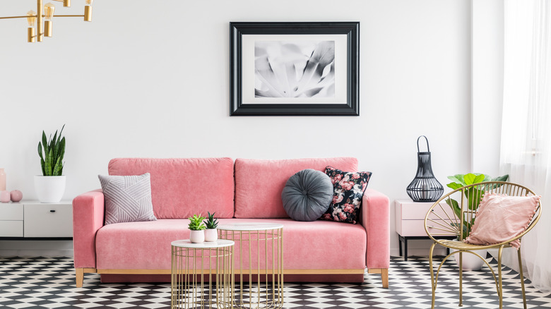 A modern living room with a black and white checkered floor, pink velvet sofa, and gold accents. The walls are white and a black and white painting hangs behind the sofa.