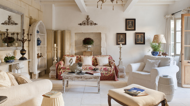 A large white living room in shabby chic style. Large, oversized fabric chairs and a dloral-covered sofa sit under a rustic chandelier and painted wood beams.