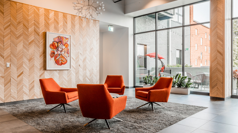 A modern apartment decorated with a vintage feel. Bright organge midcentury modern chairs are arranged in a circle around a coffee table. A parquet wood feature wall brings some warmth to white tile floors, white walls, and a wall of glass windows.