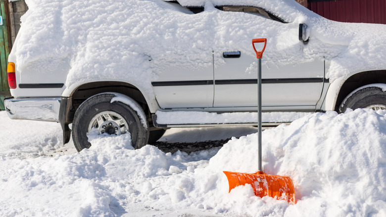 A snow-covered white car sits behind a bright, orange snow shovel.