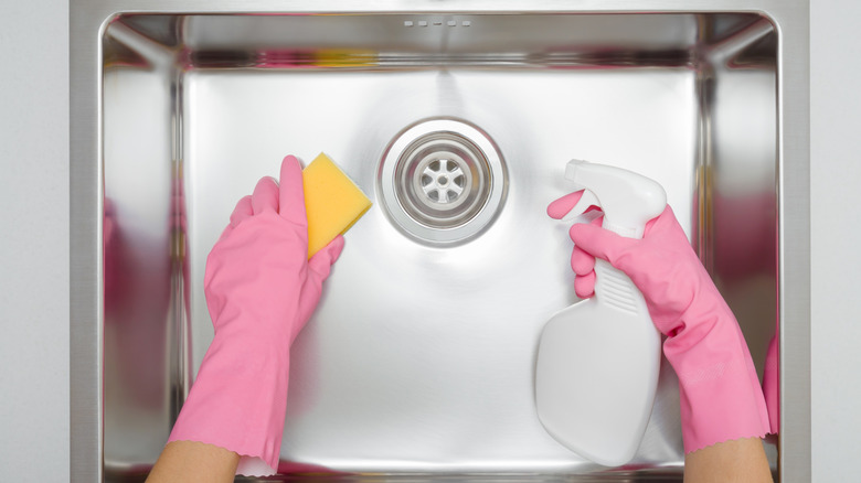 shot of someone wearing pink gloves holding a sponge and a spray bottle and cleaning a stainless steel sink