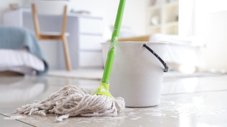 Soapy mop with a green handle resting against a white bucket. The mop is in a dorm room with tile floors.