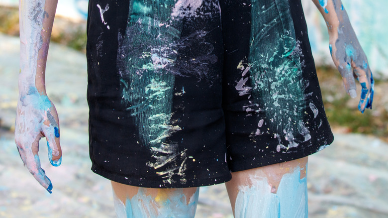 waist-to-knee shot of a woman with long red hair wearing a cut off top and shorts. Her clothes, arms, and hair are covered in paint.