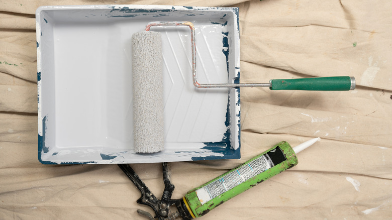 cream painters drop cloth with a green tube of caulk loaded into a caulking gun and a paint tray filled with white paint and paint roller