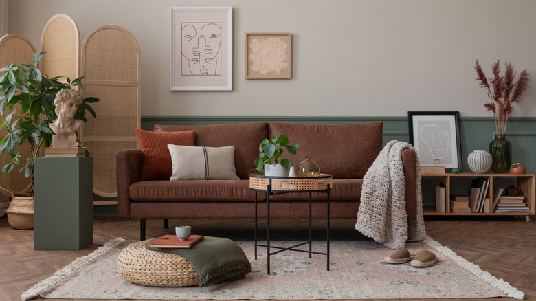 a modern bohemian living room with a rust brown sofa, muted green wainscoting, neutral medallion rug, abstract face artwork, and natural woven accents