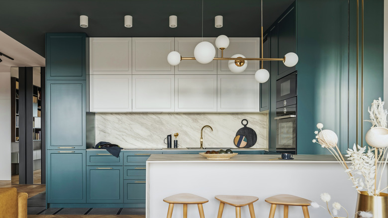 modern kitchen with dark teal lower and full height cabinets, white uppers and island cabinets, marble countertops and backsplash, and a Sputnik-style brass chandelier