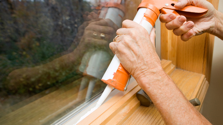 An orange and white caulking gun is used alongside a wooden windowsill.