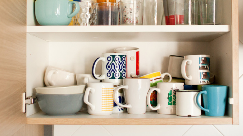 interior of open upper cabinet with an unorganized assortment of mugs stacked and piled on the lowest shelf