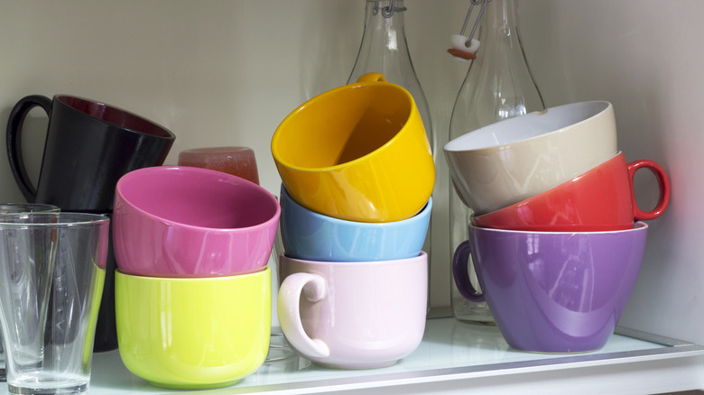 A colorful selection of coffee mugs and glasses sit haphazardly in a kitchen cabinet.