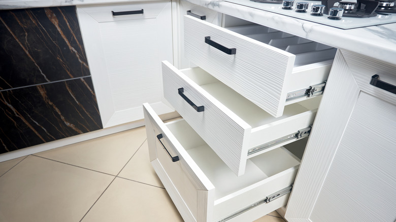 Three lower kitchen sliding drawers found under the stove top are pulled out to showcase storage potential in a white kitchen.