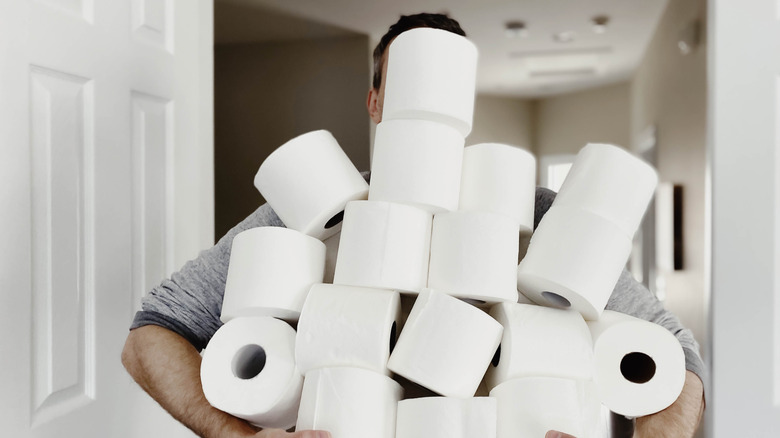 A man is walking down a hallway in a home, holding a huge pile of toilet paper rolls. His face is obscured by the rolls.