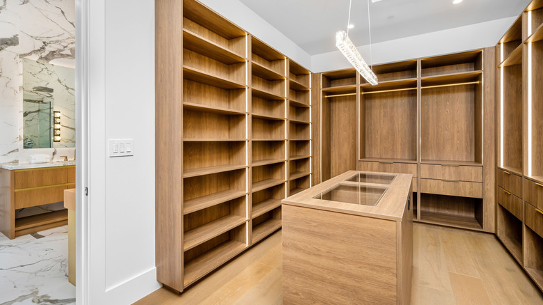 a custom wood walk in closet with an island and a door leading to a bathroom with marble look floor to ceiling tile and wood vanity