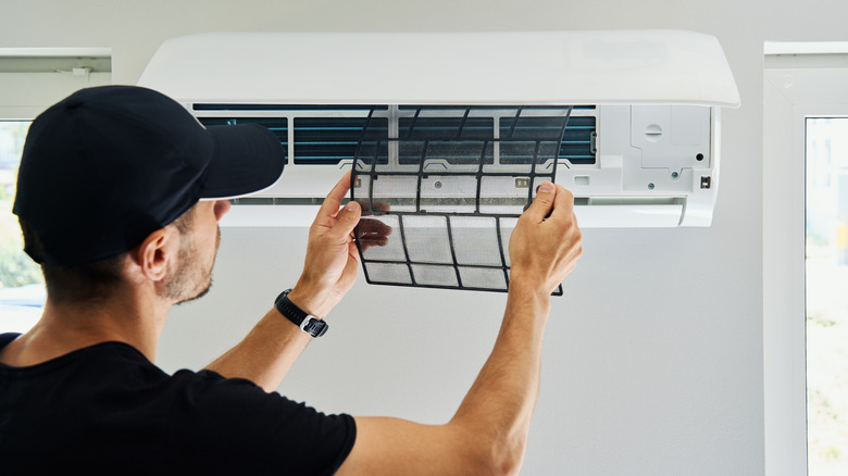 A man in a black baseball hat and black shirt is standing in front of a wall AC unit, holding up a dirty air filter.