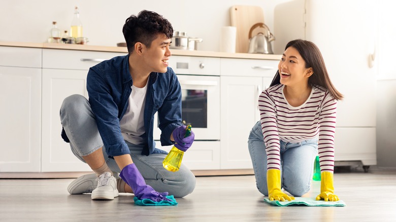 A young Asian couple is smiling at one another while they scrub the kitchen floor.