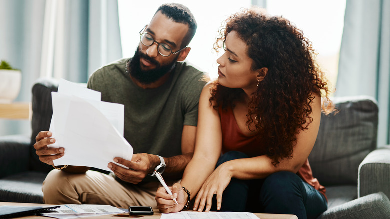 A young couple is sitting on the couch, looking through a pile of papers and taking notes.