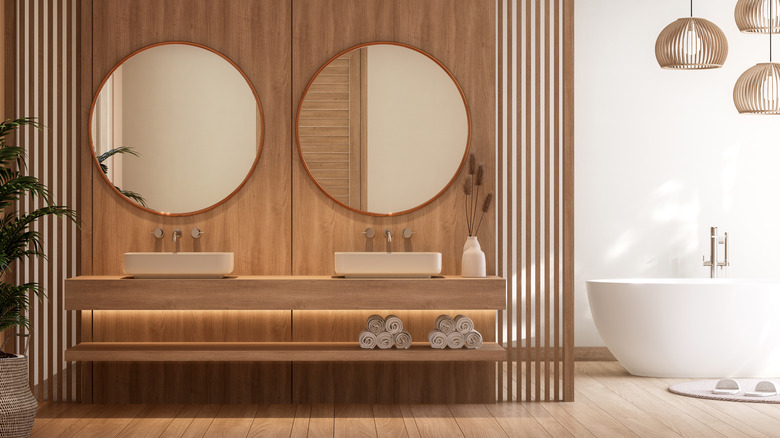 modern bathroom with natural wood paneled walls behind the vanity, floating natural wood vanity, and two white vessel sinks with wall-mounted faucets and round mirrors