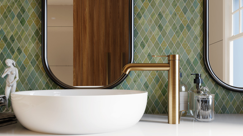 close up of a modern bathroom white vessel sink sitting on a white countertop with a side-mounted gold faucet, varied green diamond tile on the wall behind it, and oblong mirrors mounted above it with a reflection of a natural wood linen cabinet