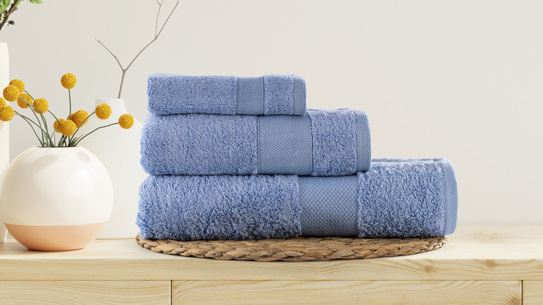 A blue washcloth is folded on top of a blue hand towel which is folded on top of a blue bath towel which is on a shelf next to a yellow fake plant