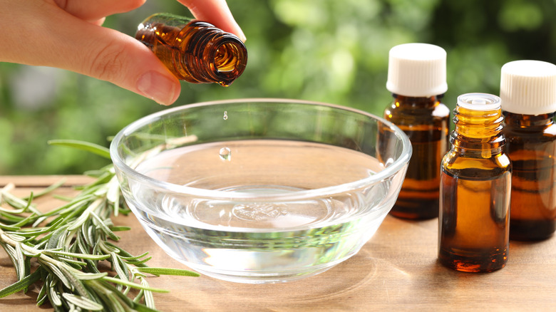 A woman's hand is adding a few drops f essential owl to a bowl of water. There are a few essential oil bottles and a sprig of rosemary on the table.